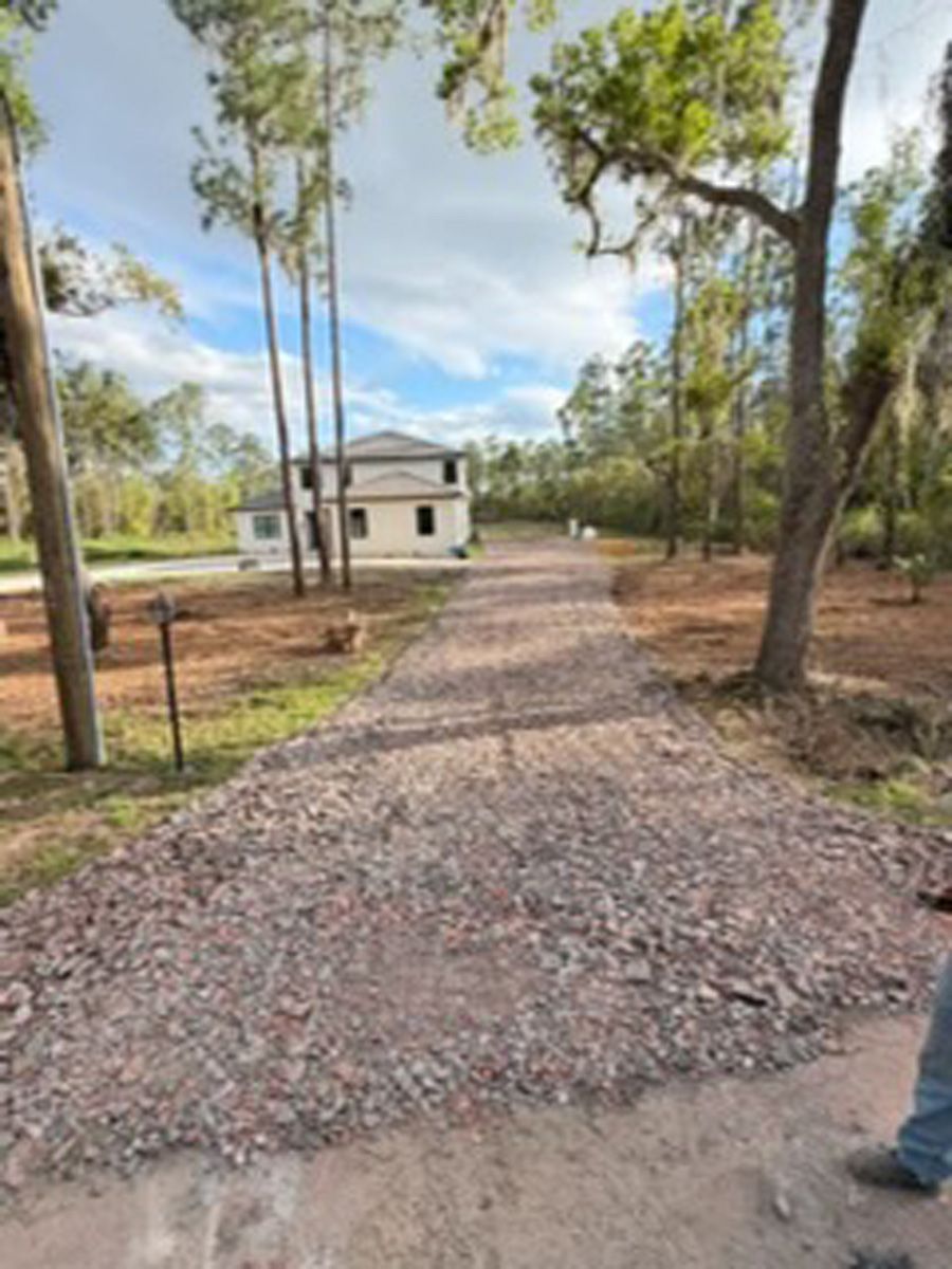 Gravel driveway leading to a small white house amid trees under a blue sky