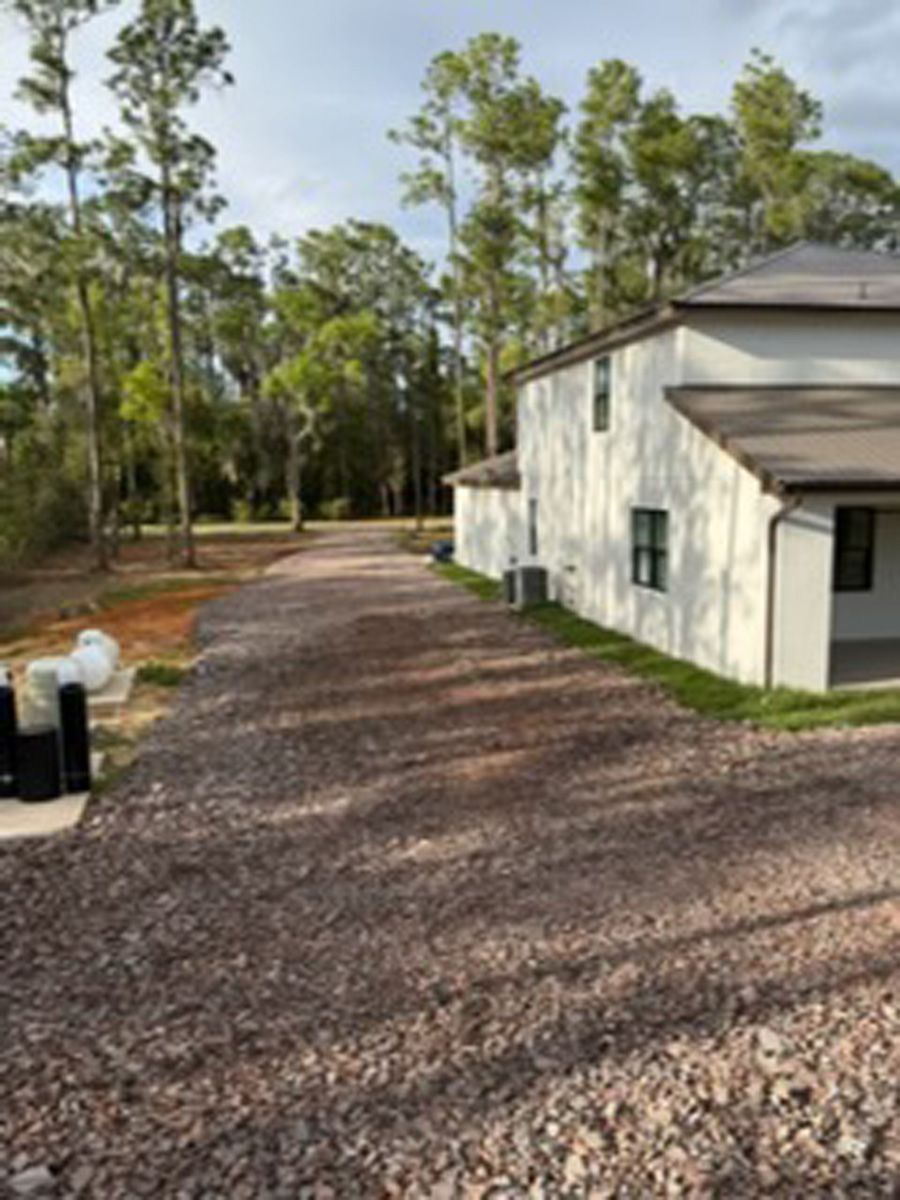 Gravel driveway beside a white house with pine trees in the background