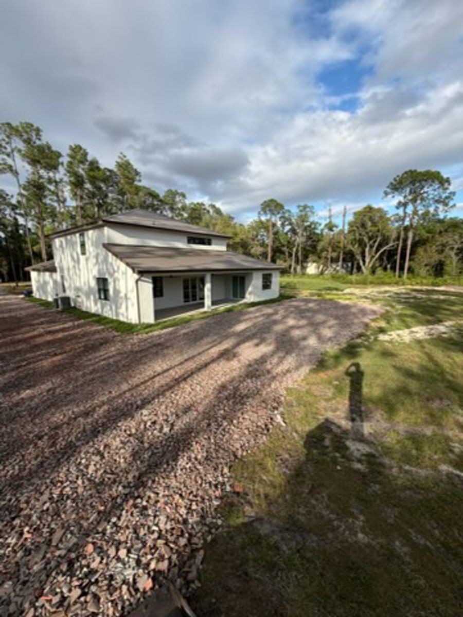White house with a gravel driveway, surrounded by trees under a cloudy sky