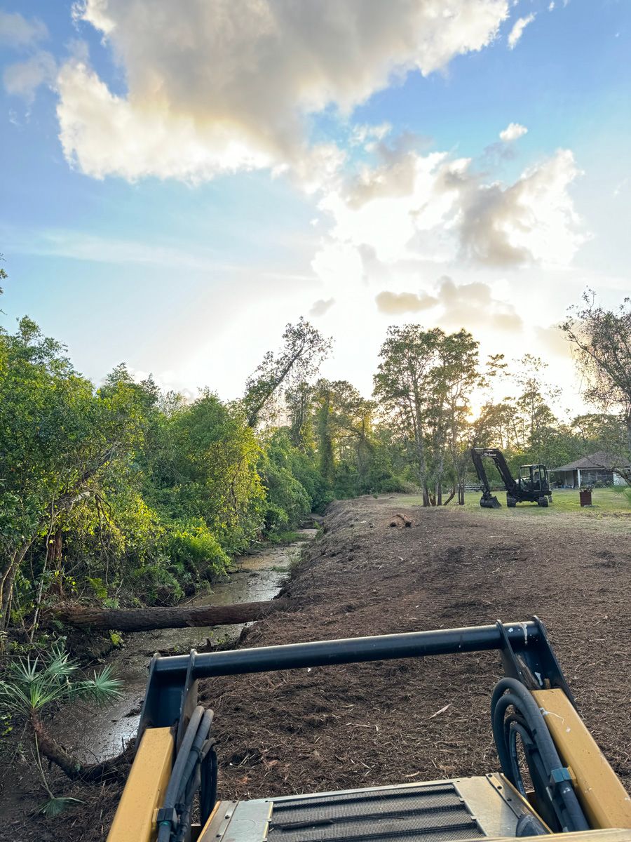 Front loader bucket on a dirt path beside trees under a bright blue sky with clouds