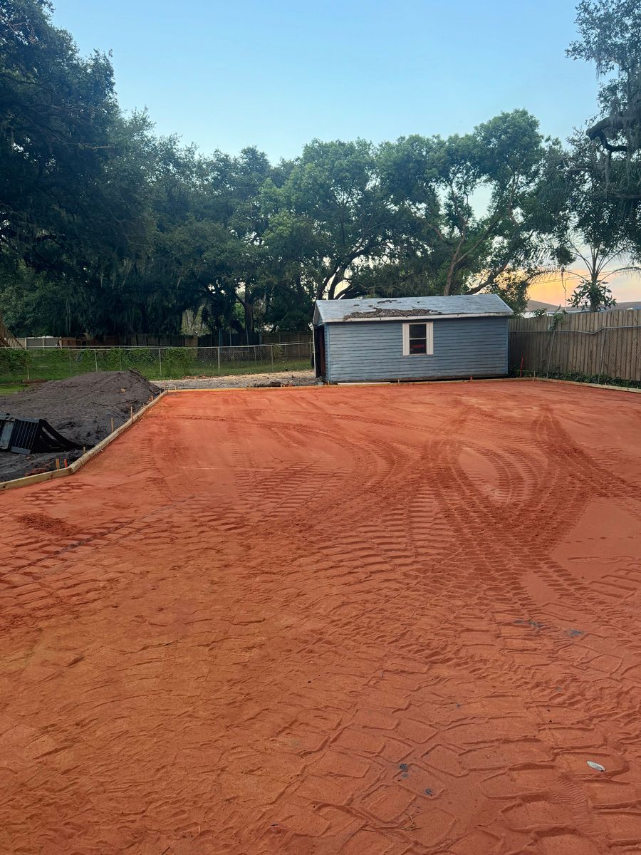 Red dirt yard with tire tracks, a small gray shed, and trees along the fence line.