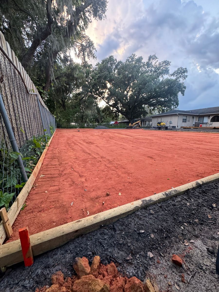 Freshly graded red dirt lot bordered by wooden beams, with a stone wall and trees nearby.