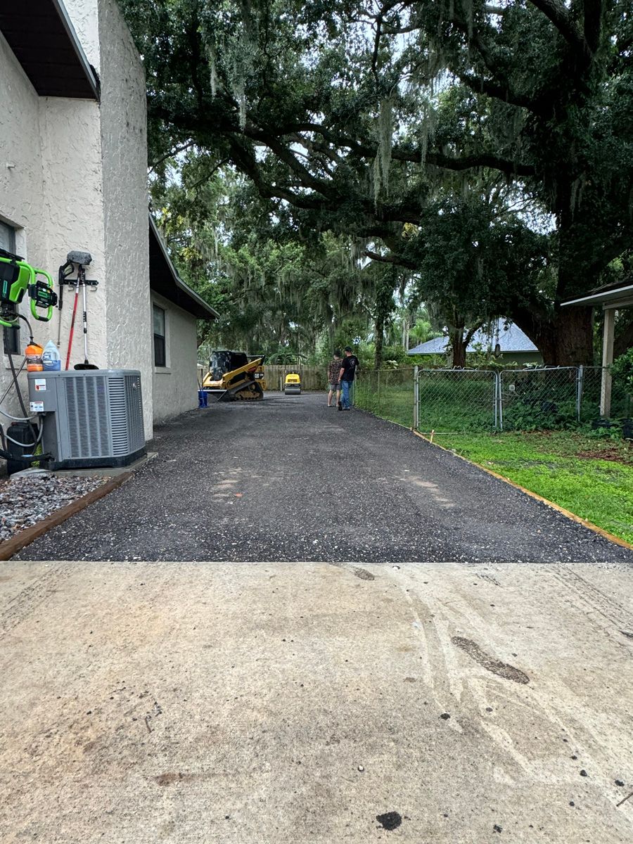 Driveway beside a house with a gravel section, large trees overhead, and a person in the distance.