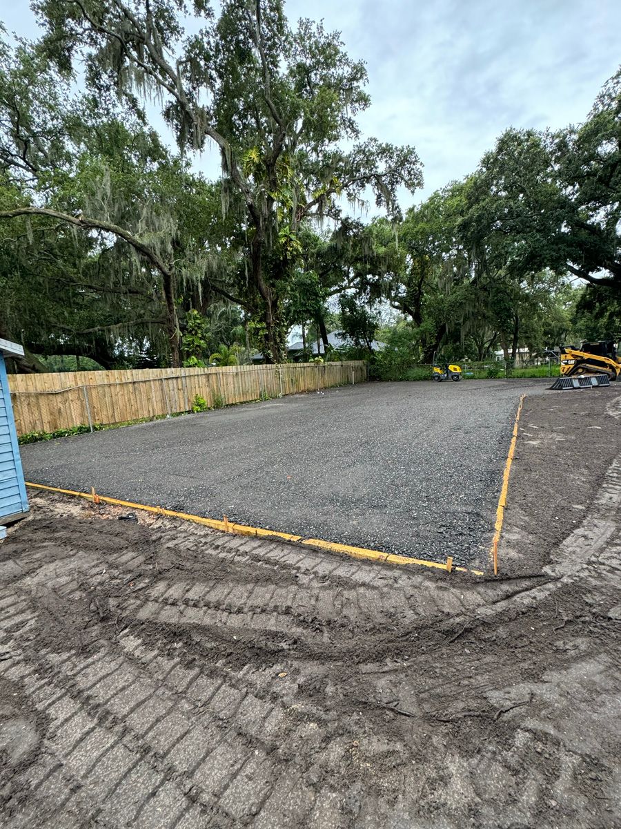 Freshly paved gravel lot bordered by trees and a wooden fence, with a small excavator at right.