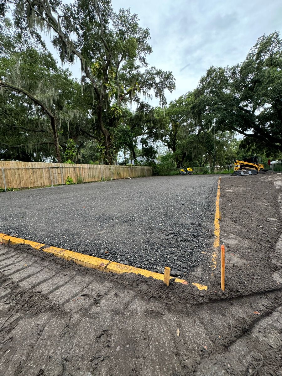 Gravel parking lot with yellow curb markings, surrounded by trees and a small excavator in the distance