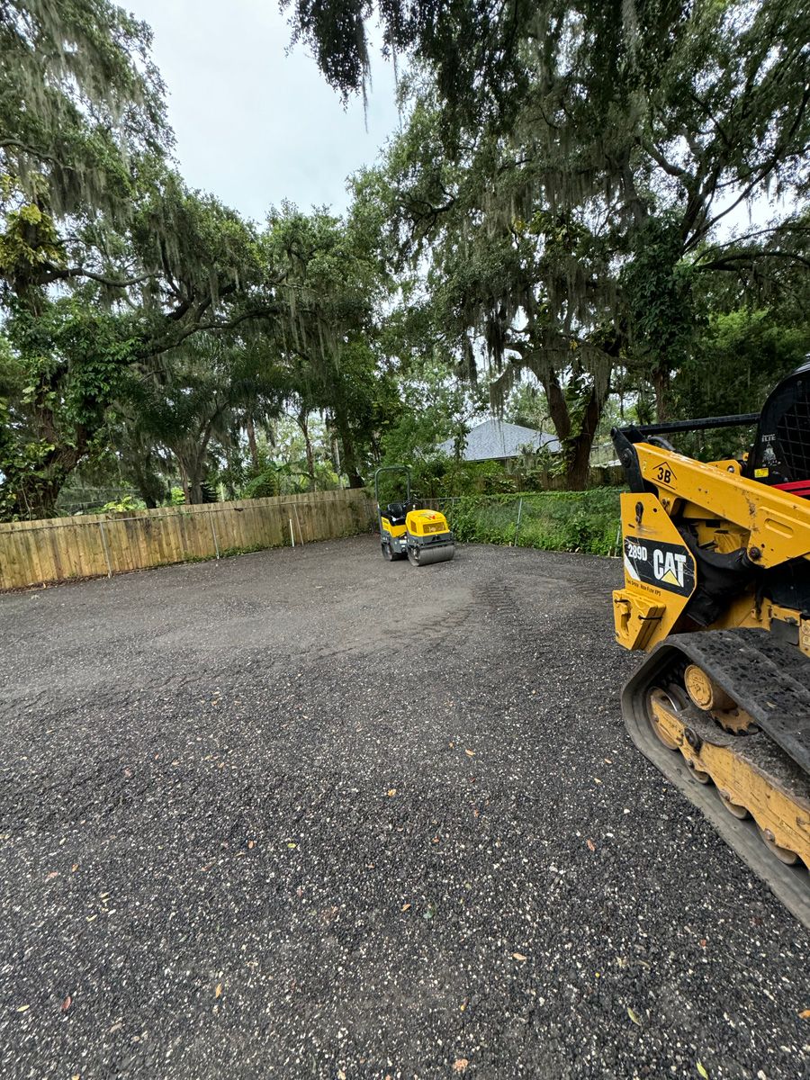 Gravel lot with trees and a yellow excavator at right, plus a small machine in the distance.