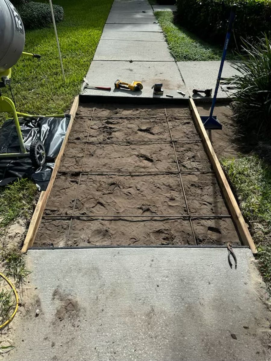 Freshly dug rectangular path section framed with wood beside a concrete sidewalk, tools at the far end.