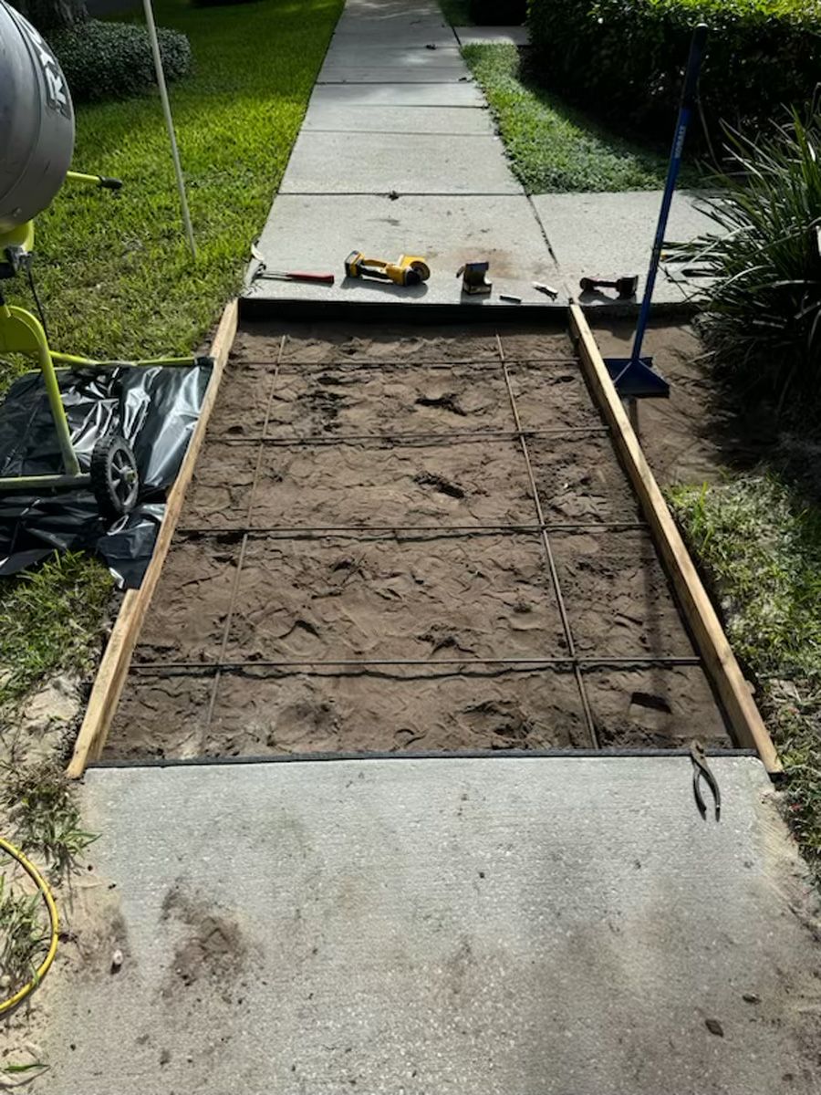 Freshly filled rectangular garden bed framed in wood beside a concrete sidewalk, with tools and trash bags nearby