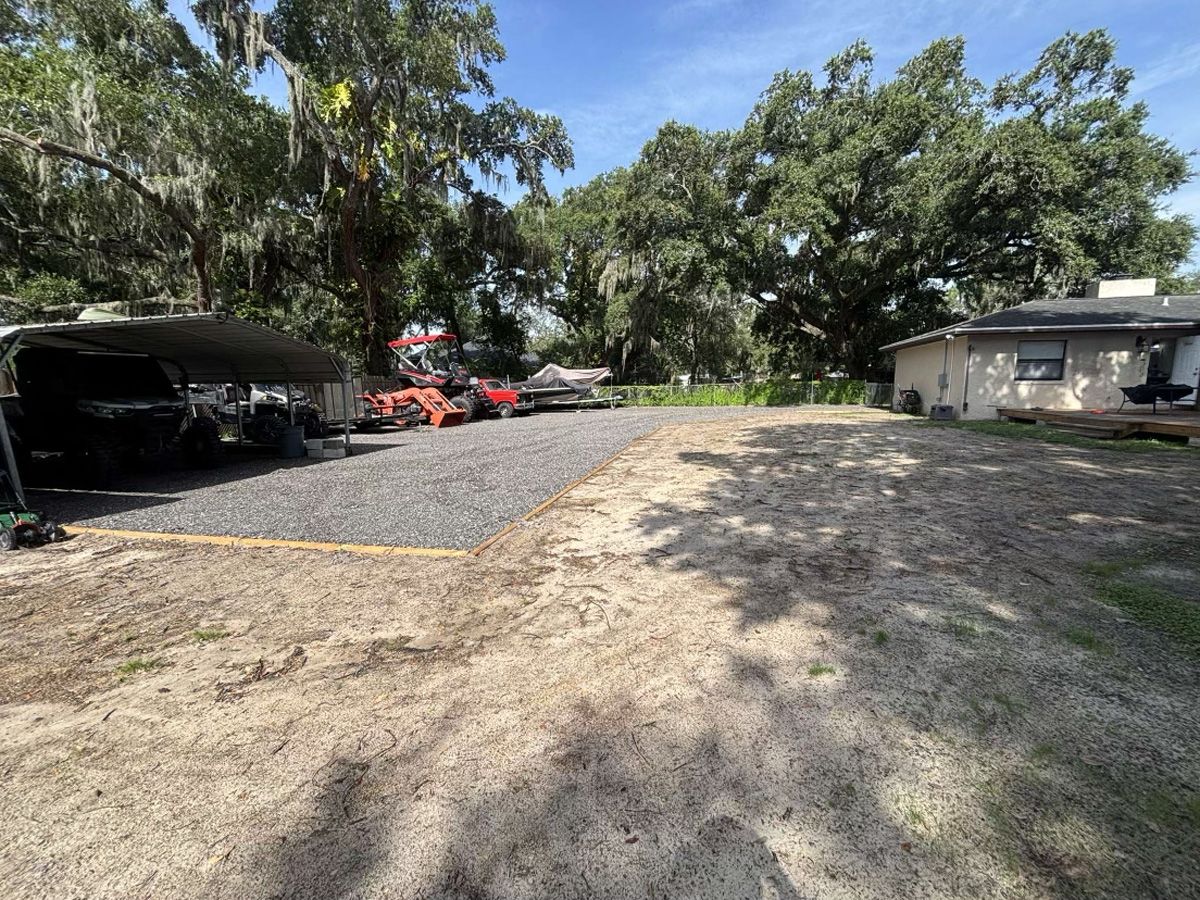Gravel driveway beside a carport and house, with trees and parked vehicles on a sunny day