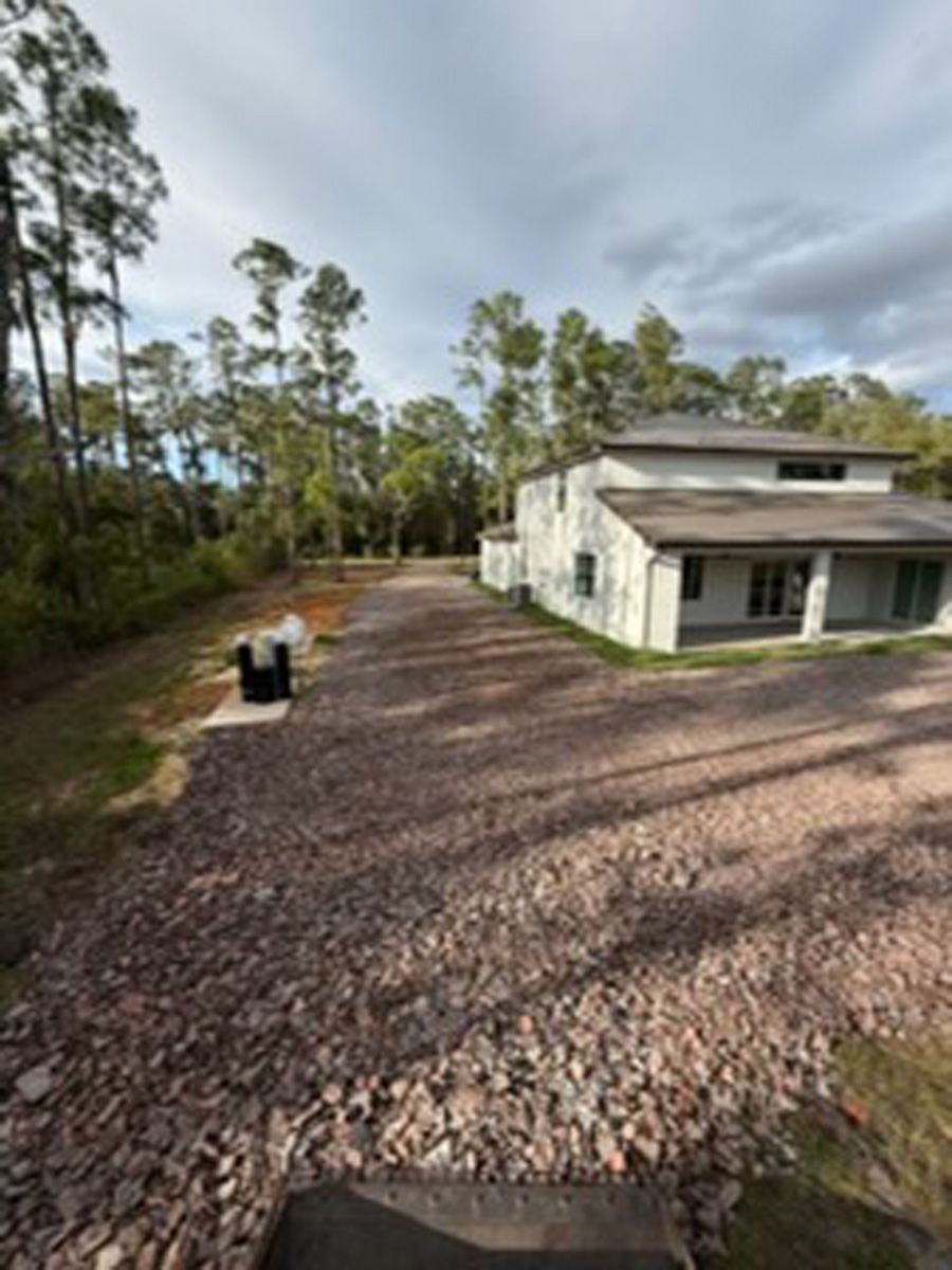 Gravel driveway beside a white house with trees and cloudy sky