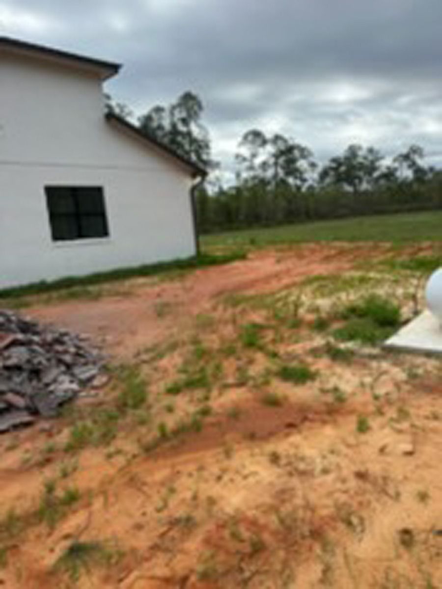 Rural yard beside a white house, with muddy red dirt, grass patches, and trees in the background