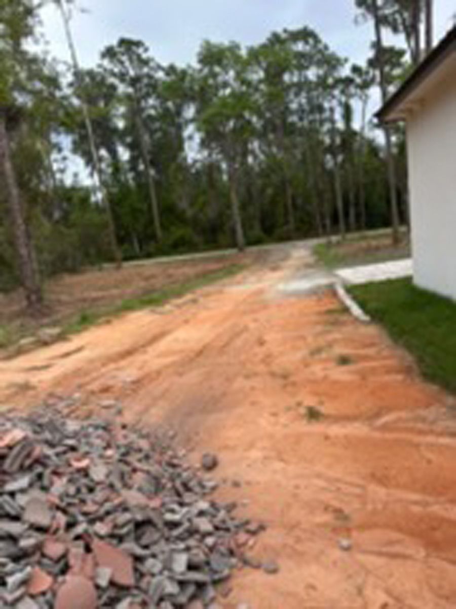Dirt path beside a white house, with a pile of broken bricks in the foreground and trees in the background