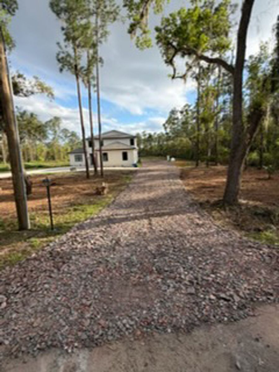 Gravel driveway leading to a small white house surrounded by trees under a partly cloudy sky