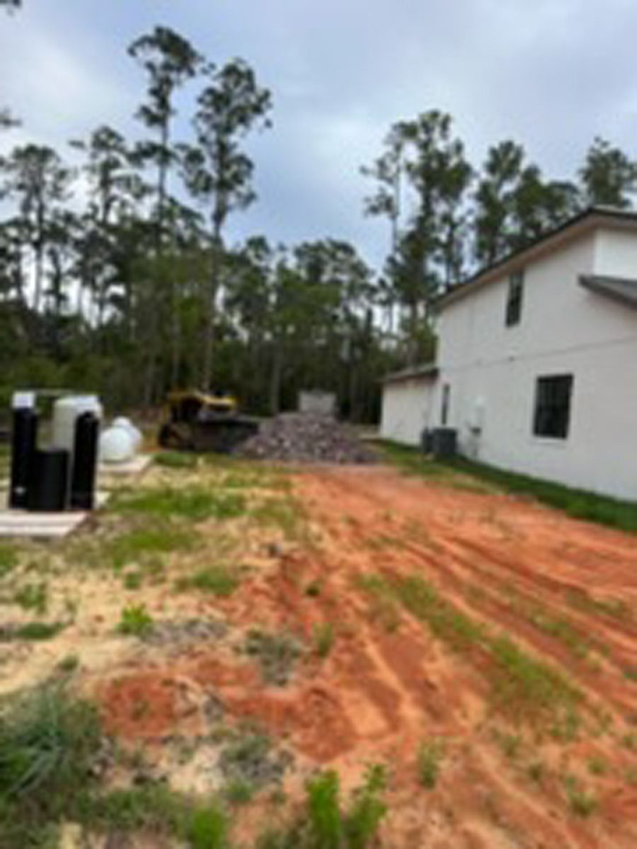 Red dirt driveway beside a white house, with construction equipment and tall pine trees in the background.