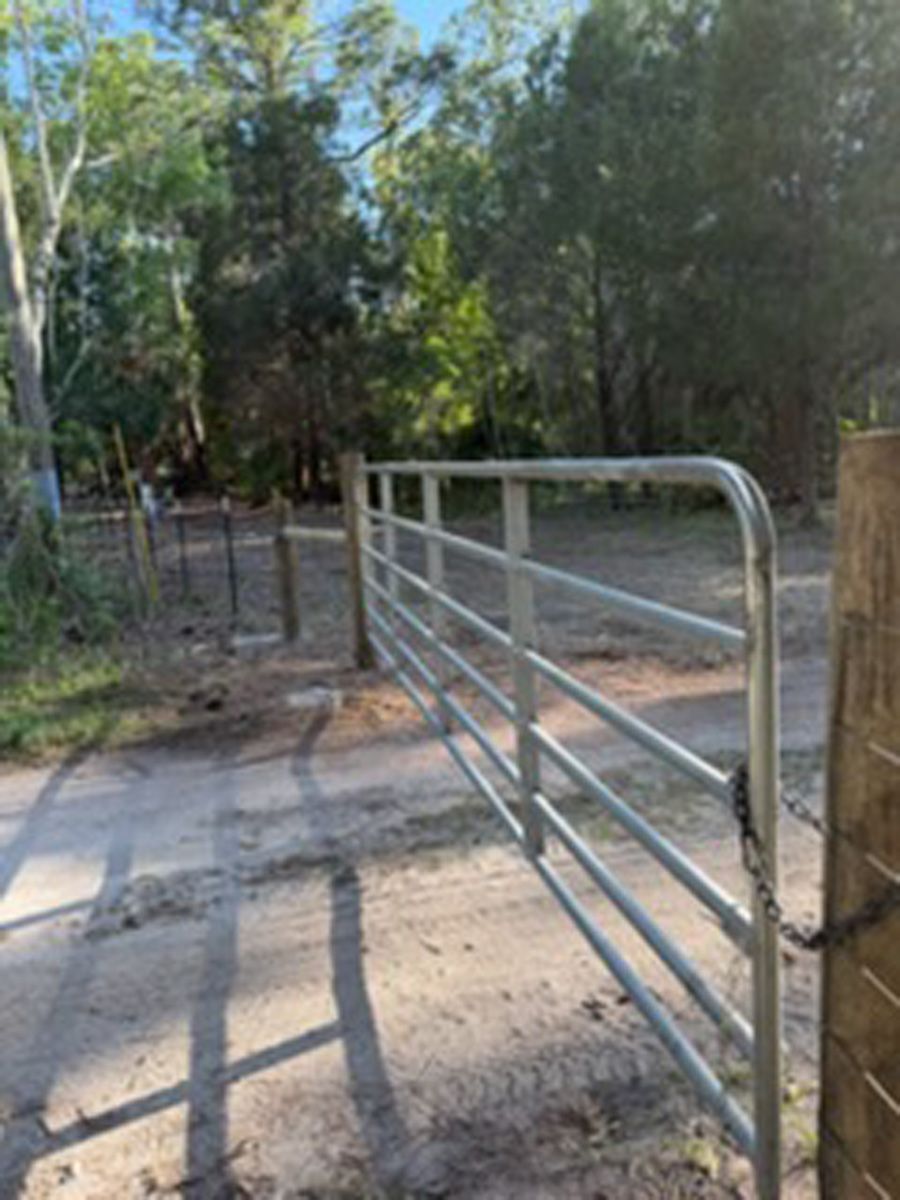 Metal gate across a dirt path in a wooded outdoor area, with fence and trees in the background