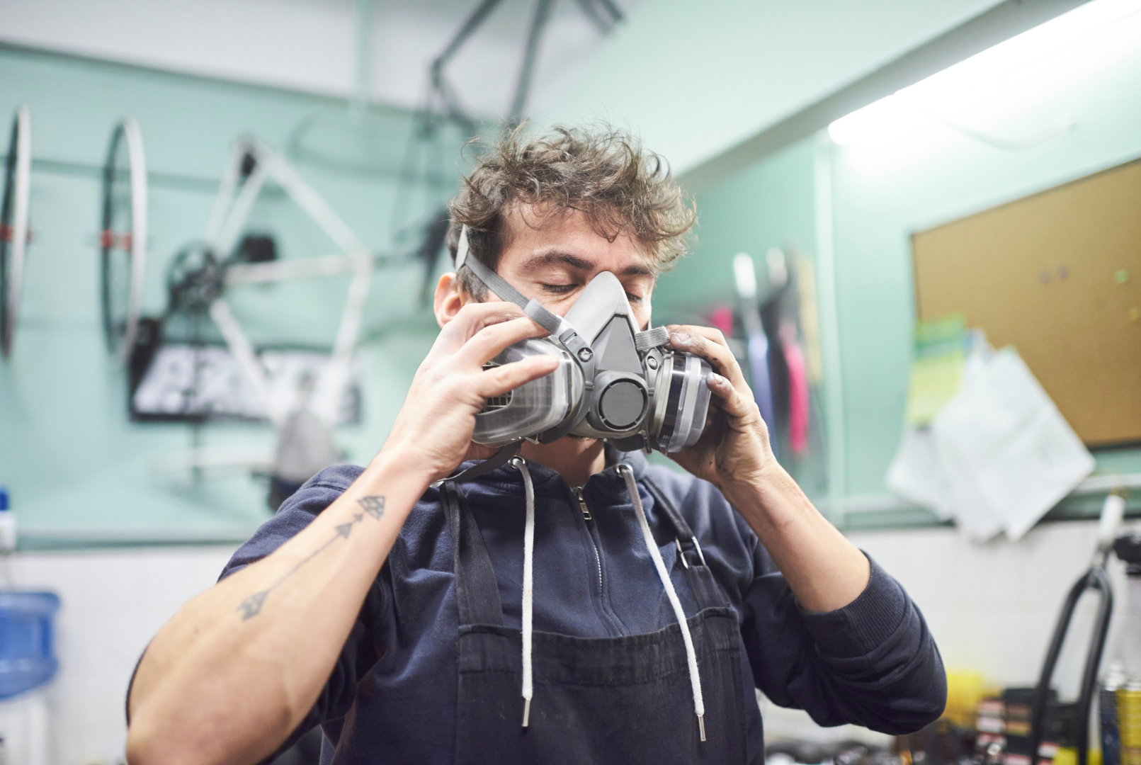 Man in workshop putting on a respirator mask; blurred bicycle and tools in background.