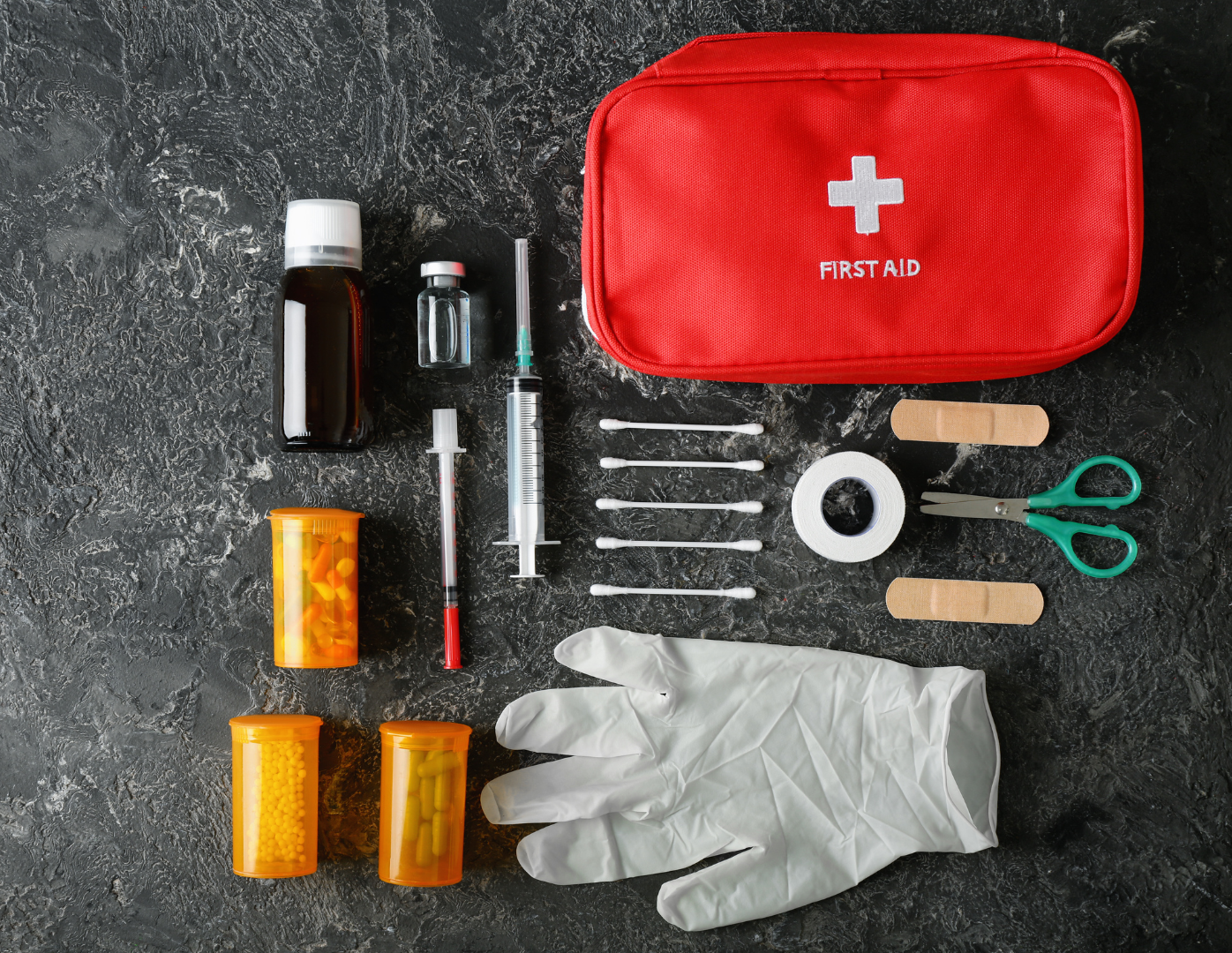 Red first aid kit with medical supplies, including medicine bottles, bandages, and a syringe on a dark surface.