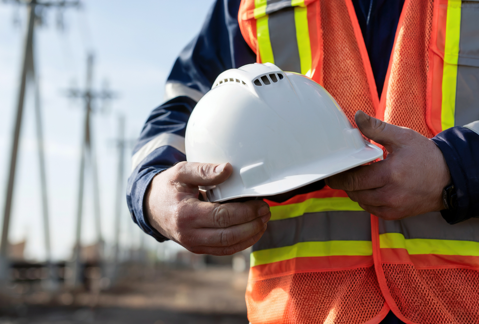 Person in reflective vest holding white hard hat near power lines.