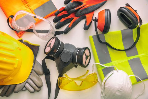 Safety equipment, including a hard hat, gloves, goggles, respirator, ear muffs, and a safety vest, arranged on a surface.