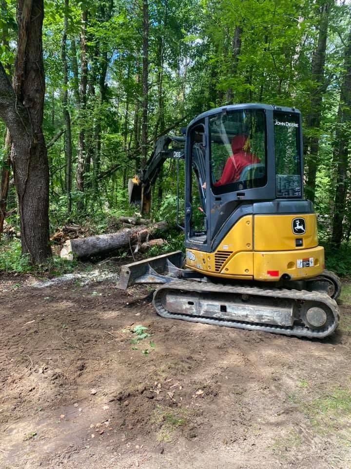 Yellow excavator parked on a dirt path in a wooded area, with a worker inside the cab.
