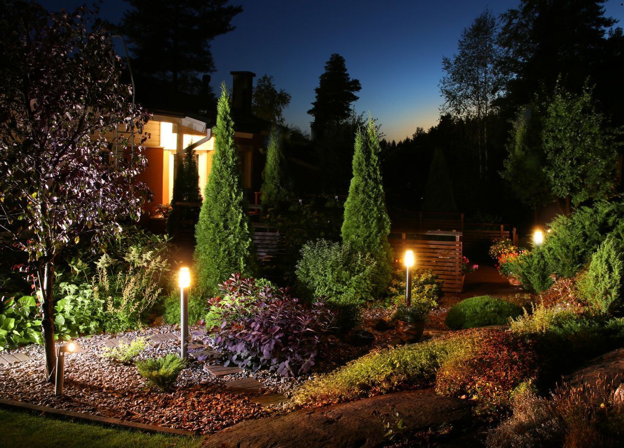 Nighttime garden illuminated by warm lights, featuring shrubs, trees, and a house in the background.