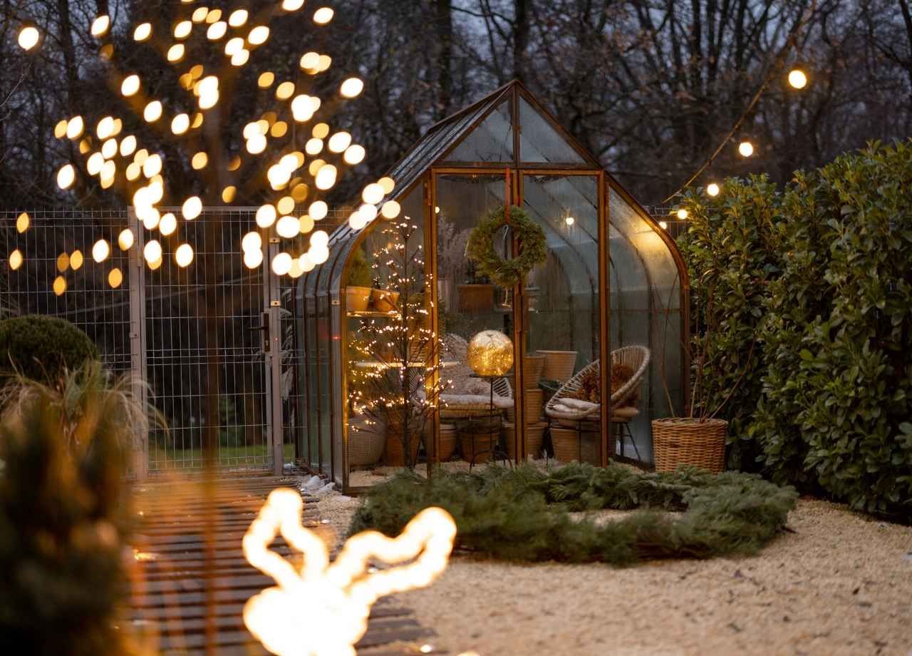 Greenhouse at dusk, lit with warm fairy lights. Cozy interior with seated chairs, plants, and a wreath.