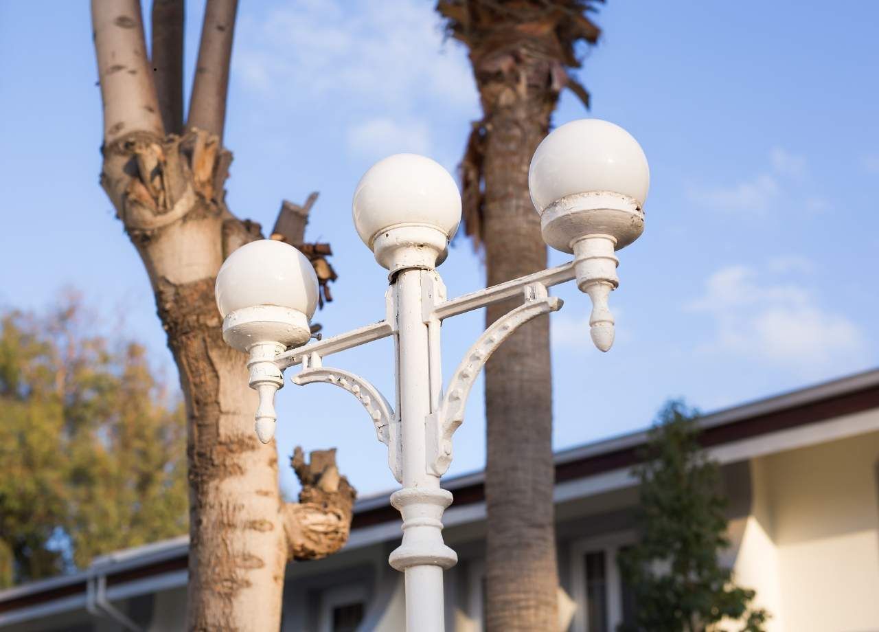 White outdoor lamp post with three globe lights, trees, and a building against a blue sky.
