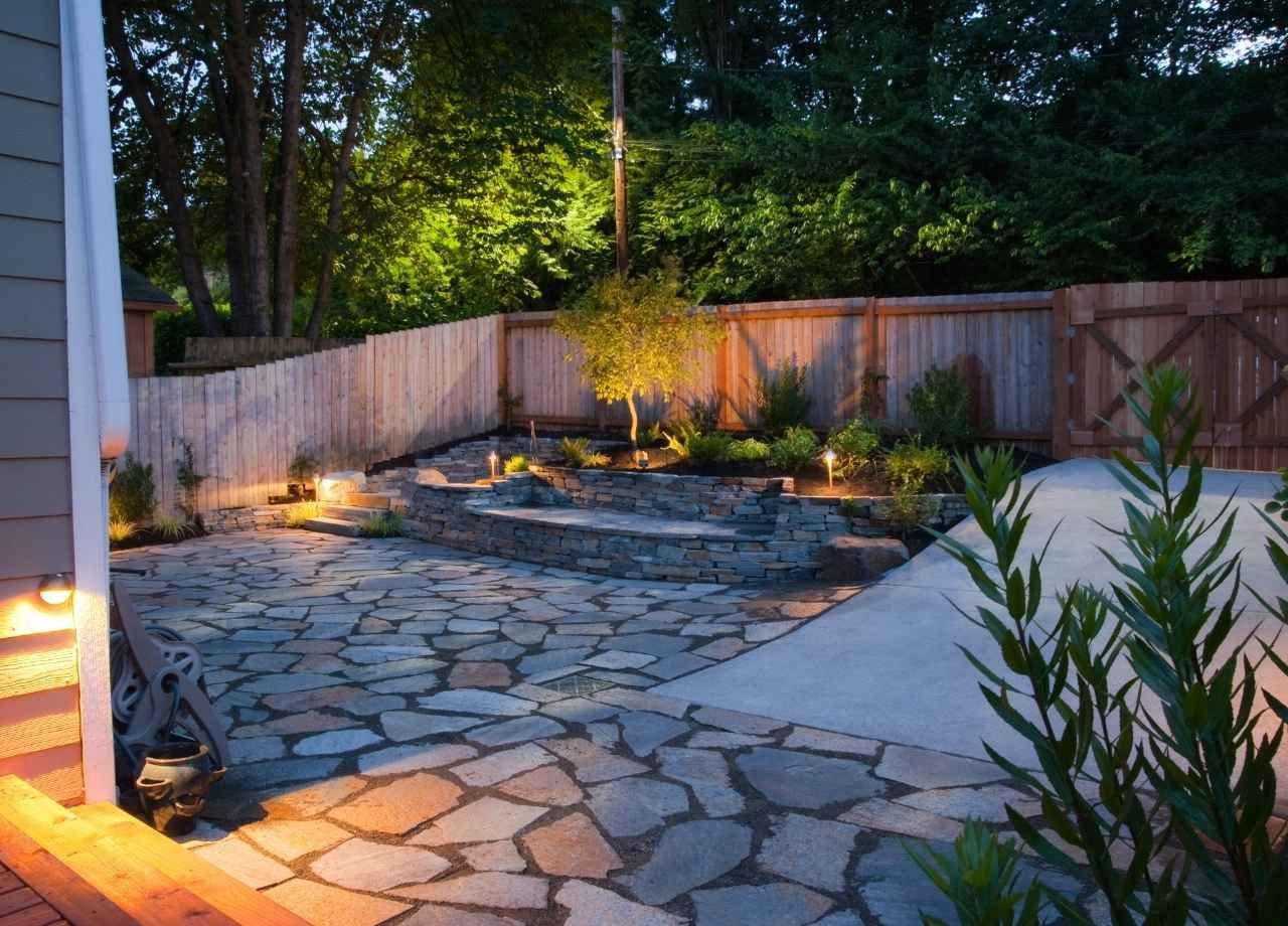 Stone patio with built-in seating and landscaping, lit by warm lights. Wooden fence in background.