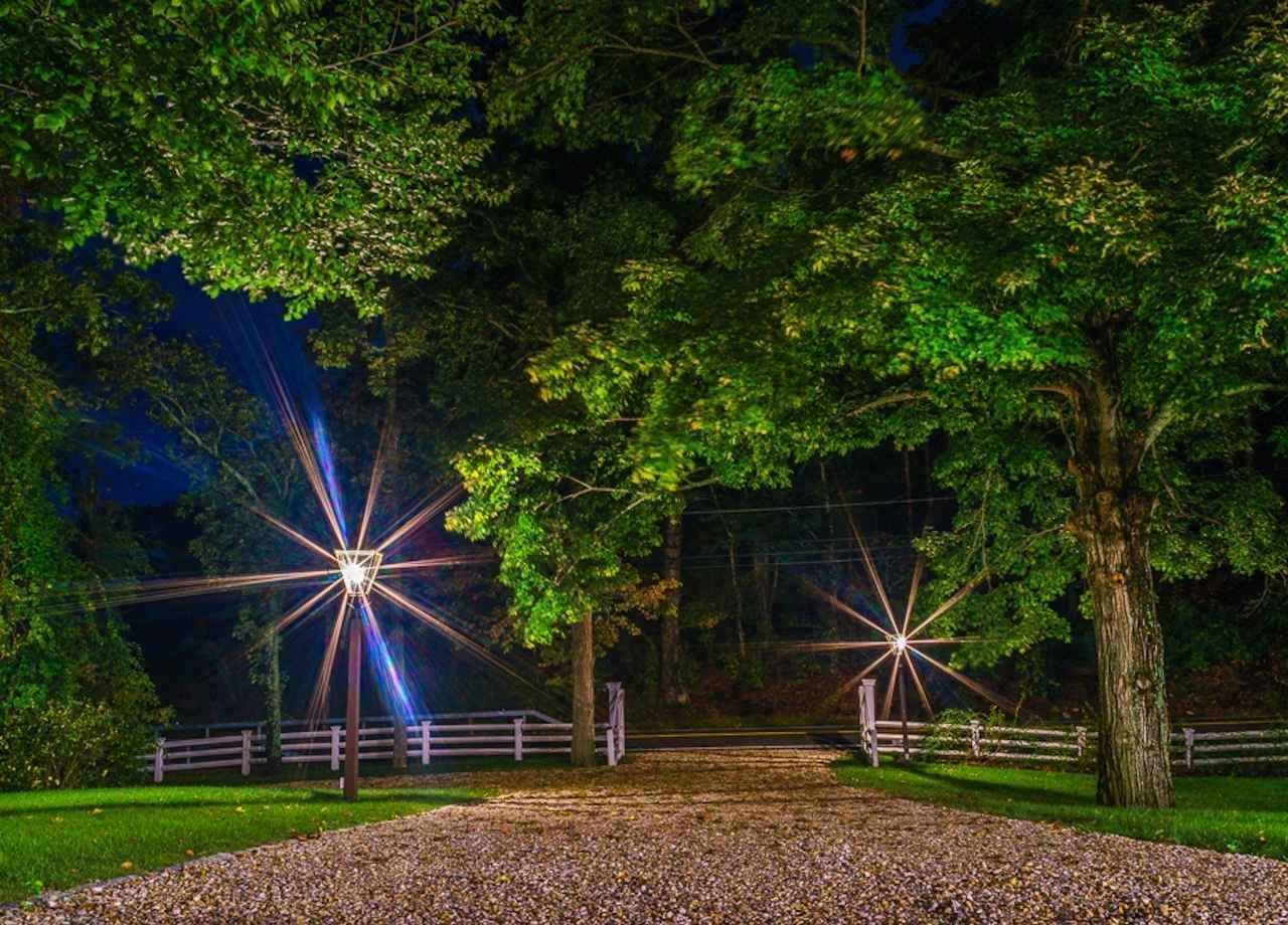 Gravel driveway leading to a white fence gate, flanked by large trees and illuminated by starburst lights at dusk.