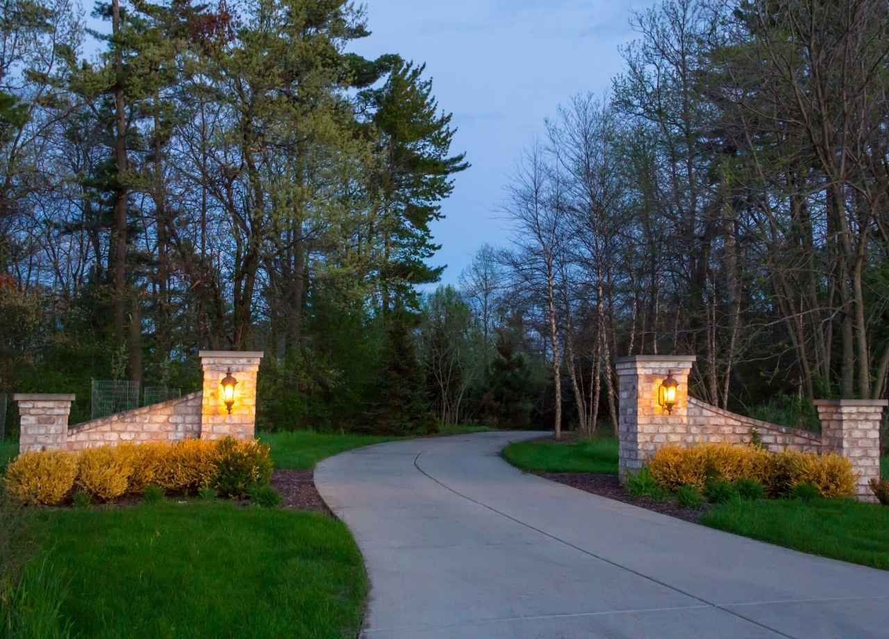 Stone entry with lit lanterns and a winding driveway, flanked by green grass and trees.