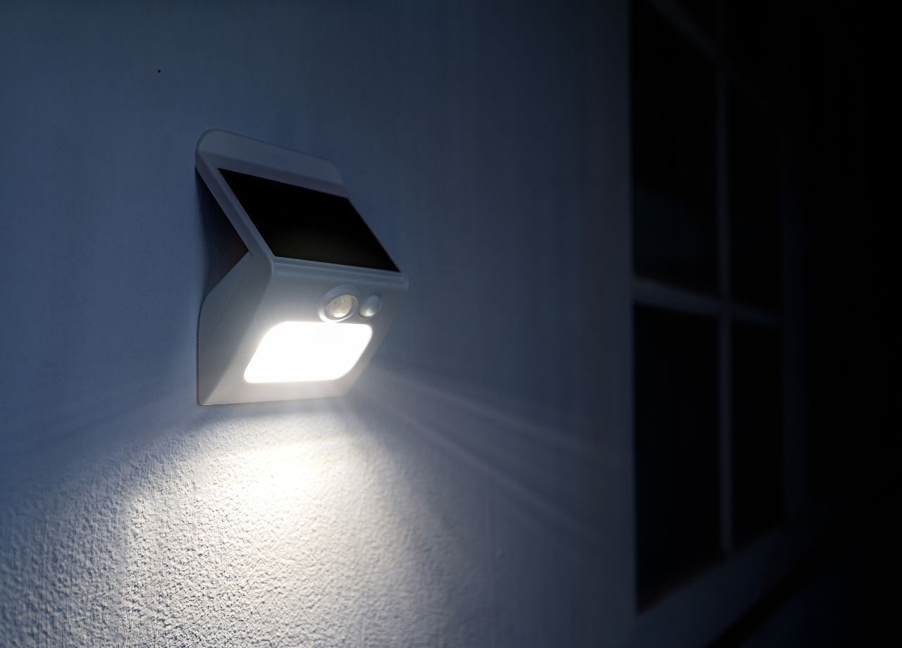 Solar-powered security light shining brightly on a white wall near a window at night.