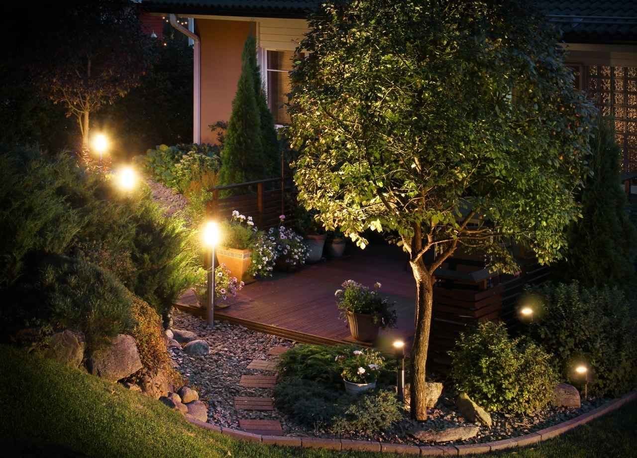 Illuminated garden path with lights, lush greenery, and a house in the background at night.