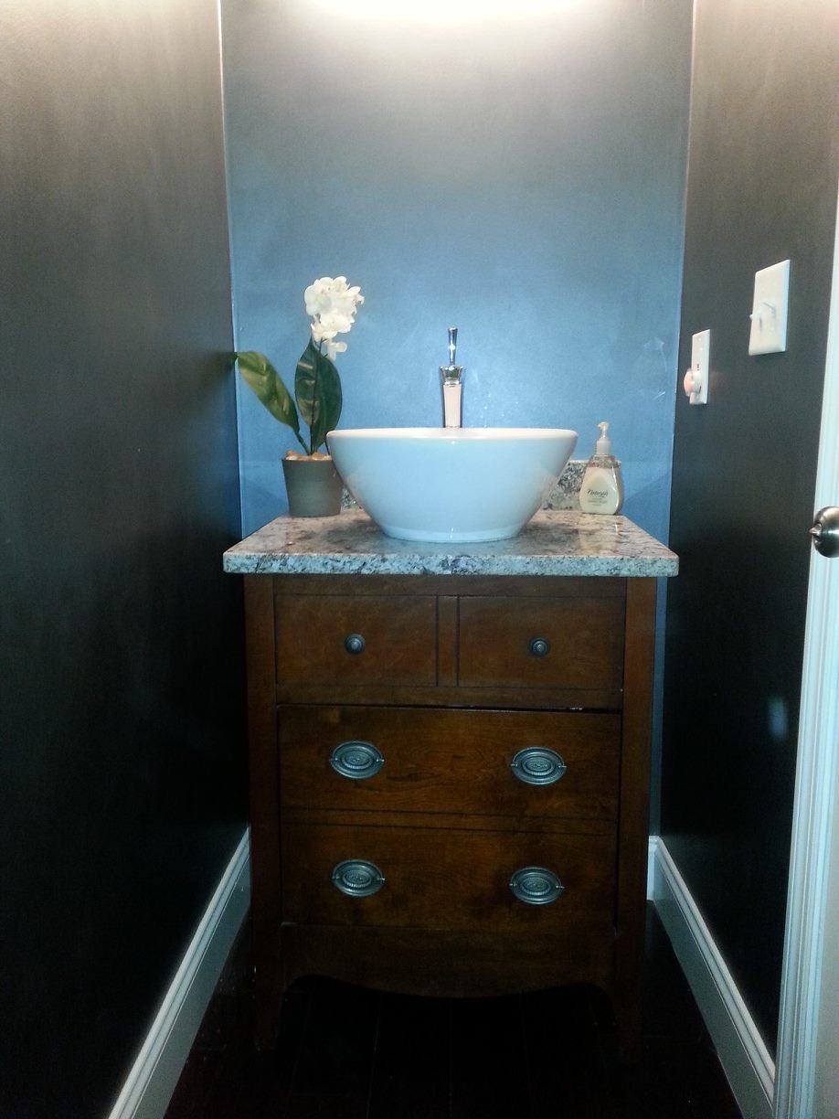Small bathroom vanity with blue vessel sink, marble countertop, and dark wood cabinet between black walls.