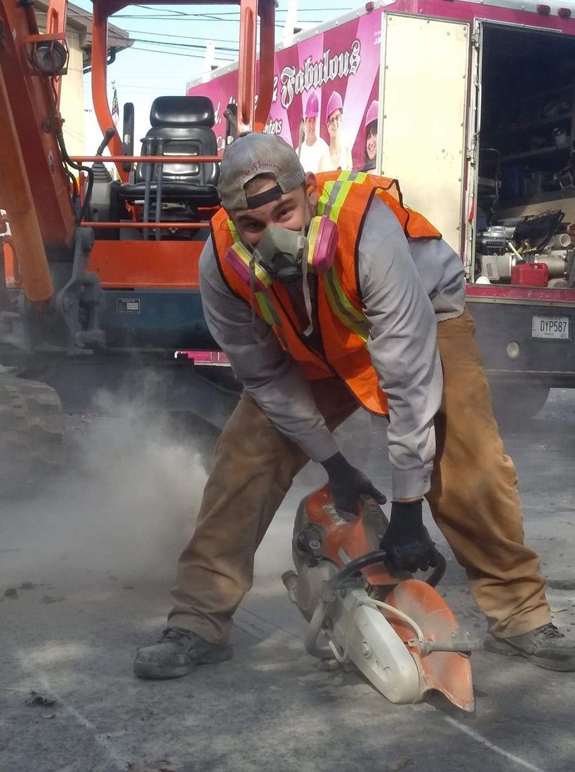 Construction worker in orange vest cutting pavement with a circular saw, dust rising around him