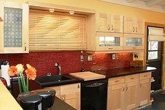 Modern kitchen with light wood cabinets, red tile backsplash, black countertops, and a window above the sink