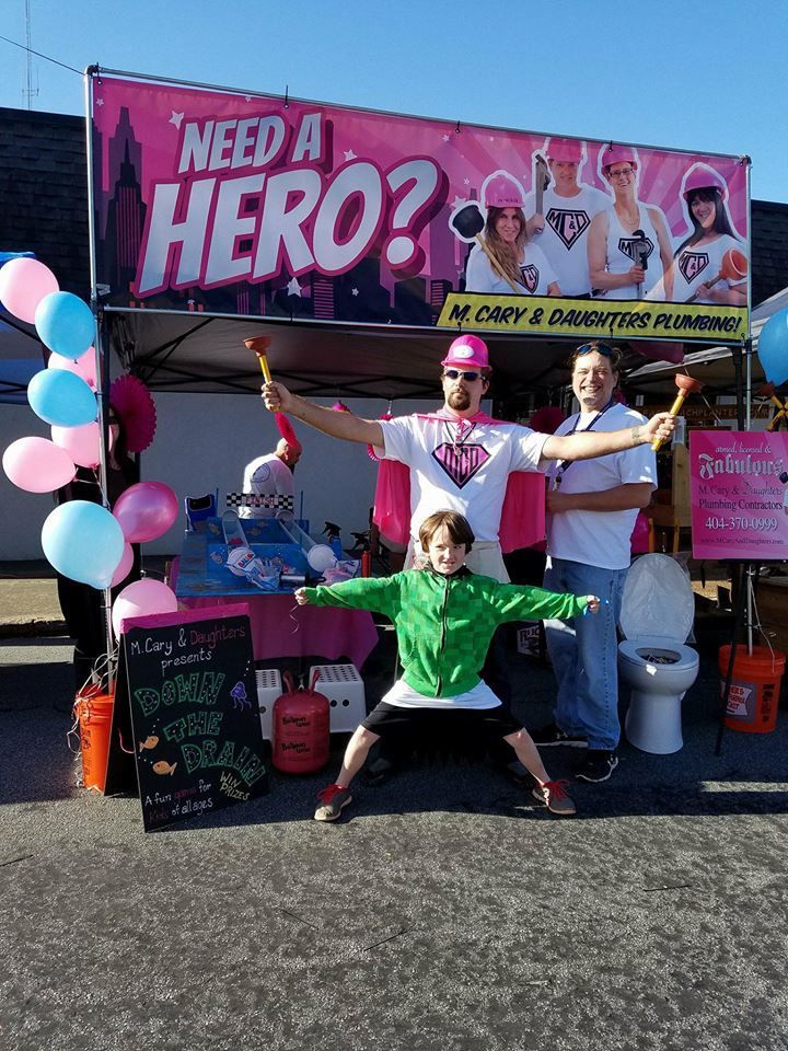 Two people holding a sign that says women at work. People posing at a pink “Need a Hero?” booth with balloons and donation signs