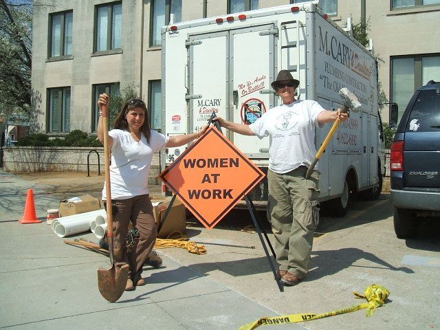 Two people holding shovels beside a “Women at Work” sign in front of a truck