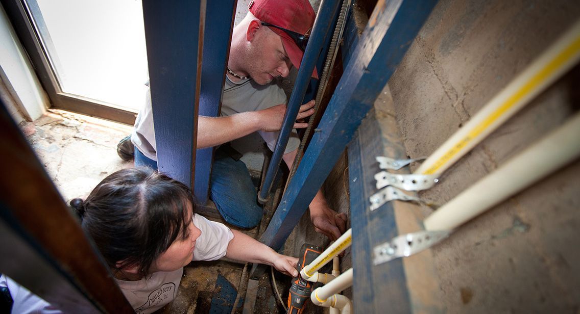 Workers crouch in a narrow trench beside blue metal beams and pipes, inspecting something below.