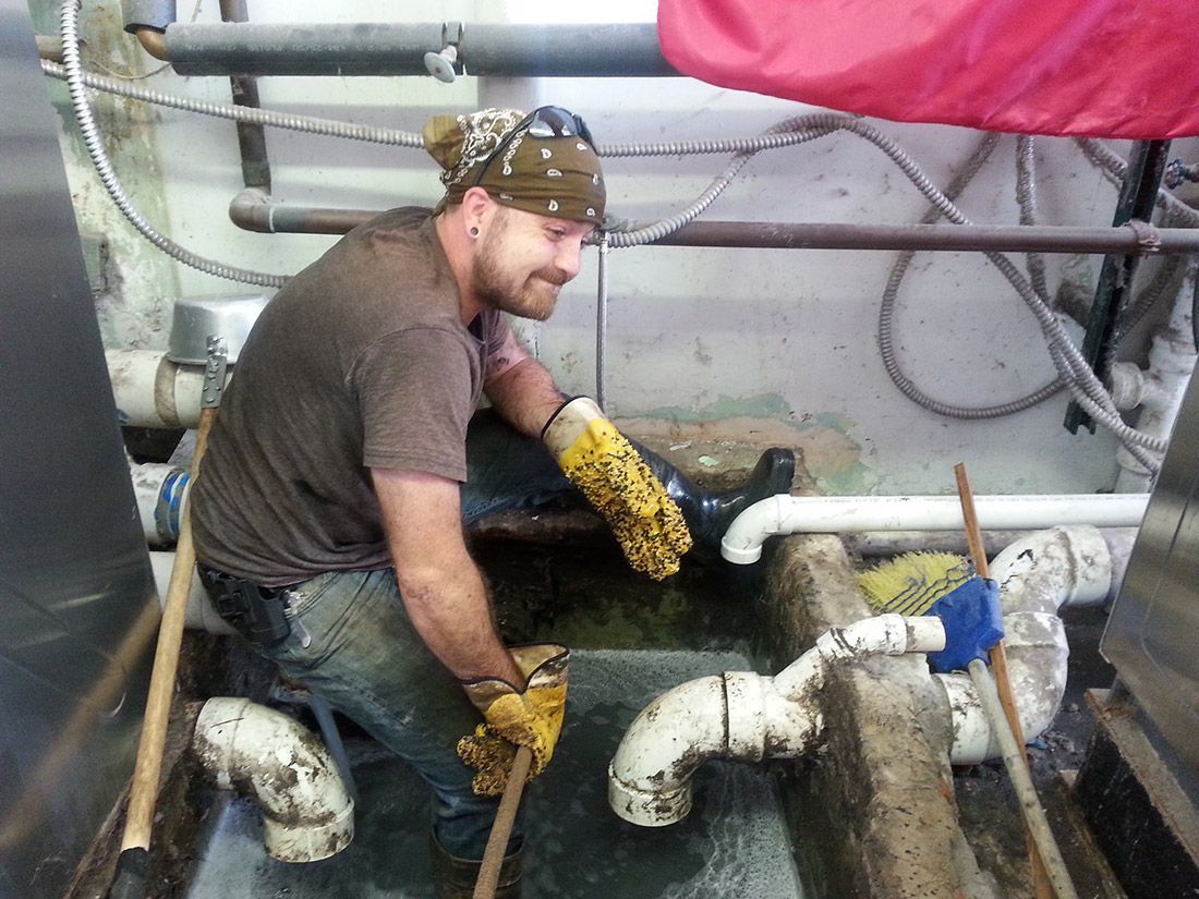 Worker kneels in a wet utility pit, handling white pipes amid industrial equipment.