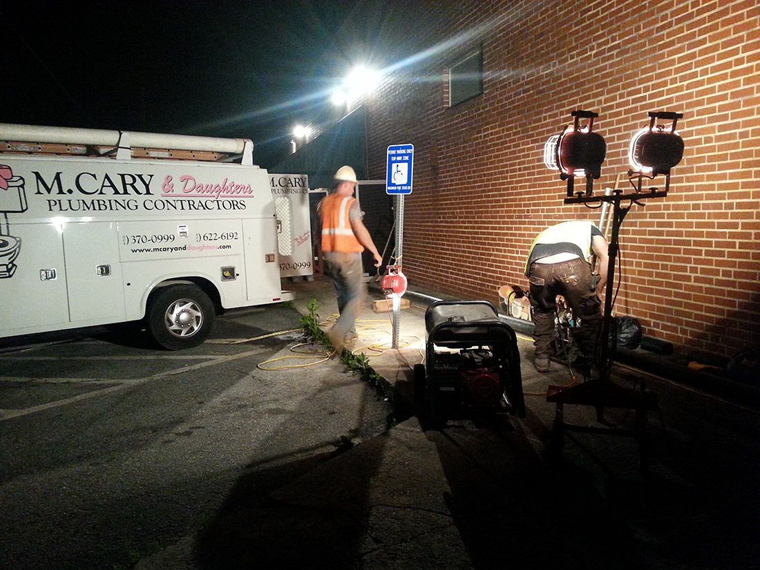 Workers and equipment beside a white moving truck at night near a brick building