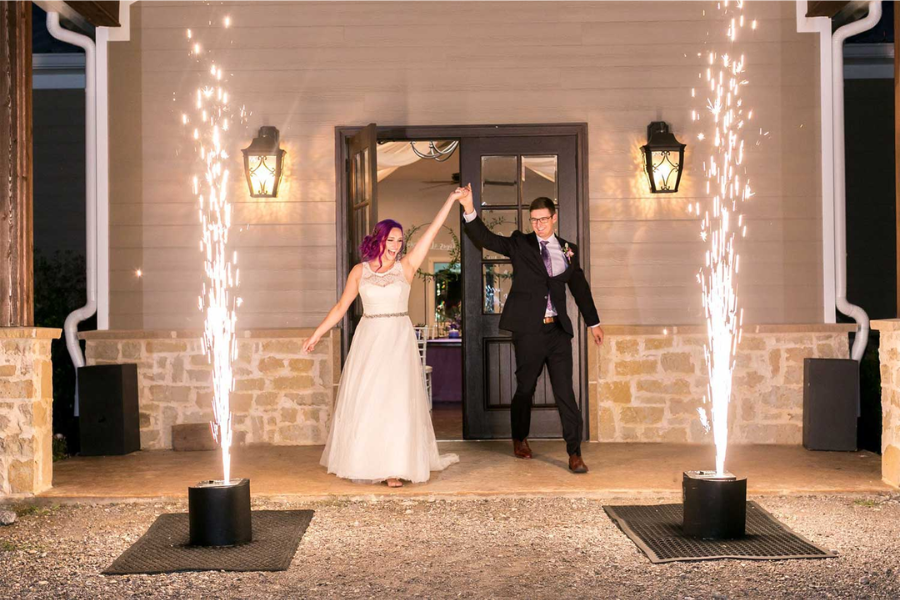 A bride and groom are standing in front of a building with fireworks.