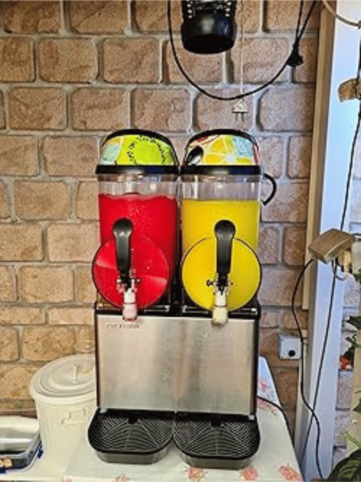 Two slushy machines are sitting on a table in front of a brick wall.
