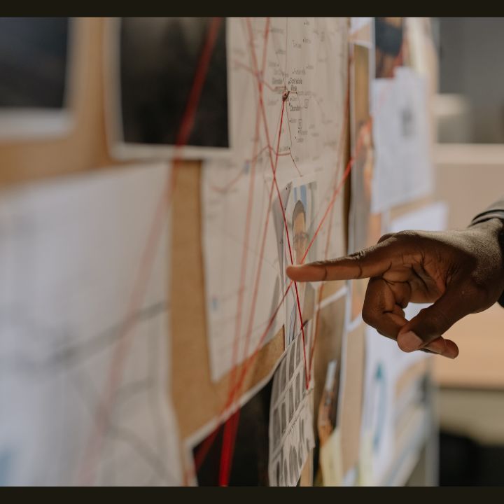 Hand pointing at a photo on a corkboard with red string connecting images and documents; an investigation scene. Private Investigator Services in La Vergne TN