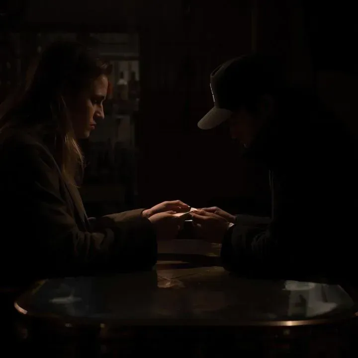 Two people, hands touching over a dark table. One wears a cap. Dimly lit interior.