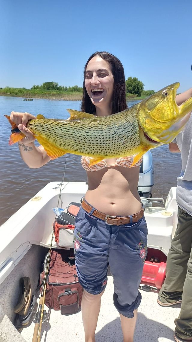 Mujer en un bote sosteniendo un gran pez dorado, sonriendo en un lago soleado.