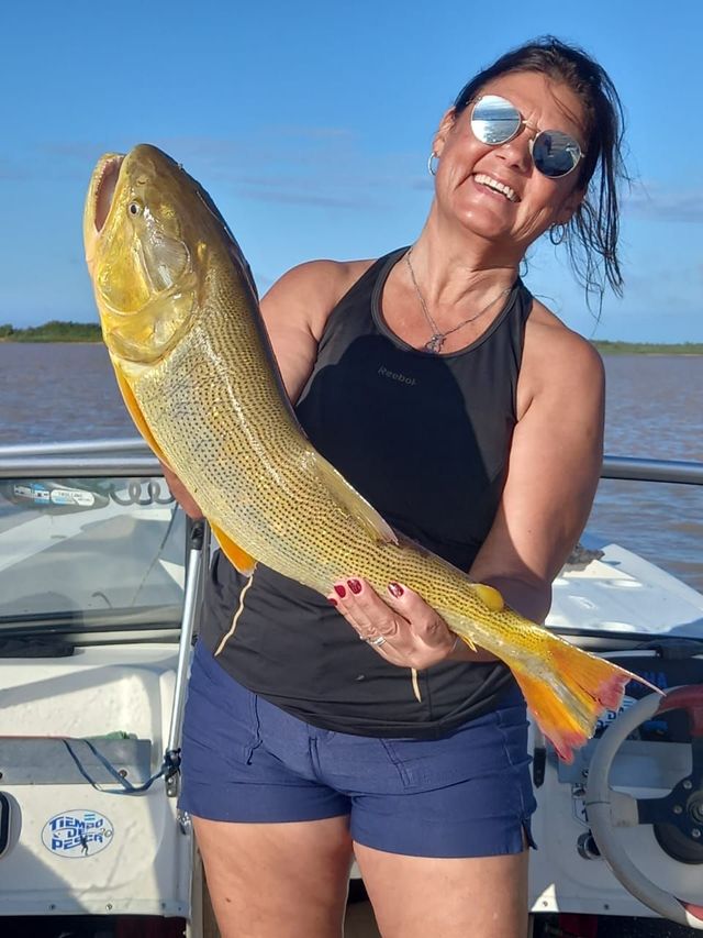Mujer en un bote sosteniendo un gran pez dorado, sonriendo con gafas de sol bajo un cielo azul despejado.