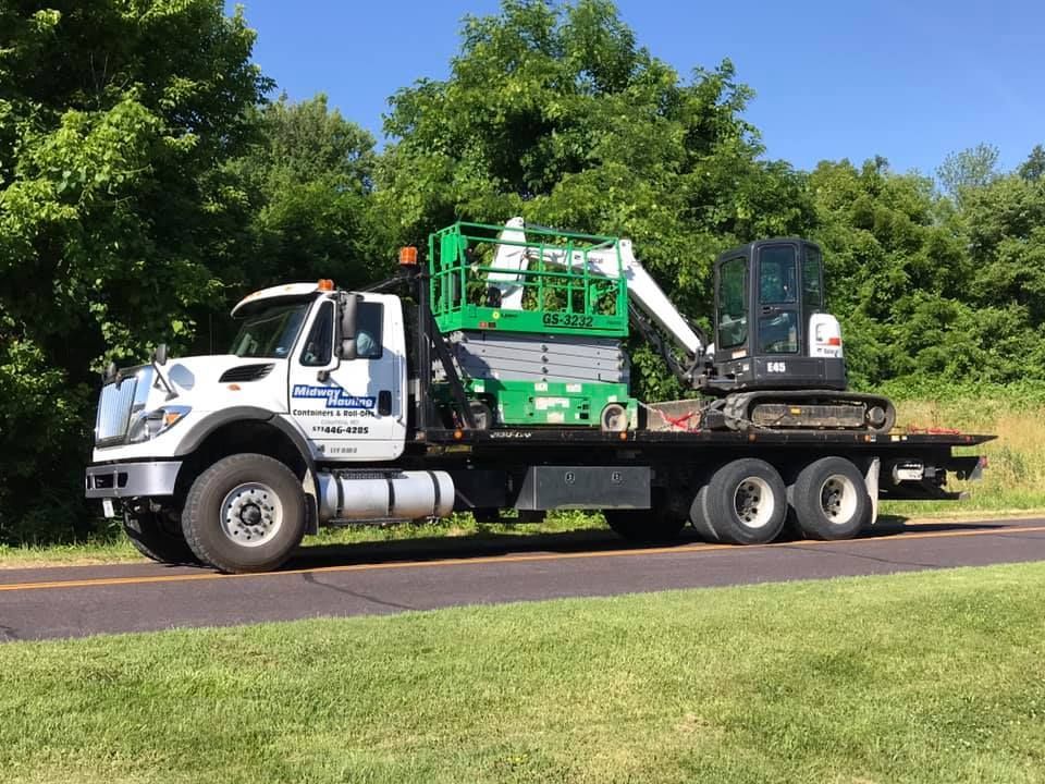 Flatbed truck carrying a green lift and small excavator on a road, with trees in the background.