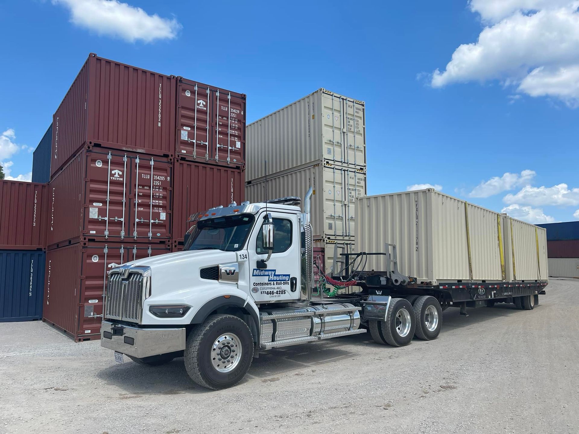 White semi-truck with trailer hauling a beige shipping container; containers stacked in background. Blue sky.