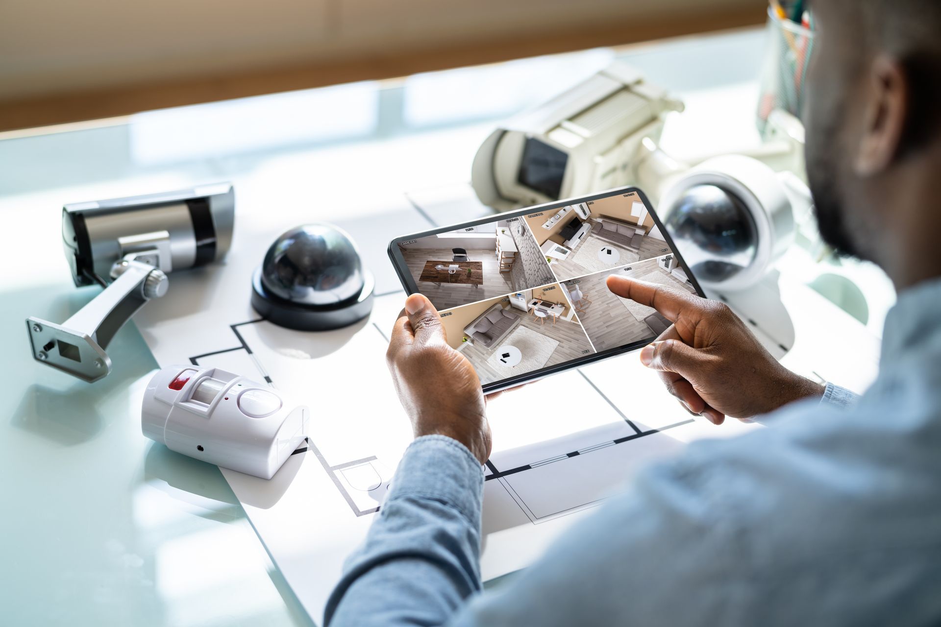A man uses a tablet to monitor security cameras, with various security cameras on his desk.