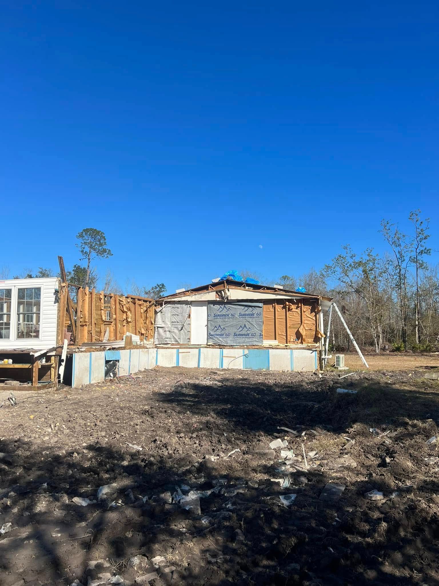 A damaged, wall-less house frame sits on a concrete foundation in a debris-strewn field under a clear blue sky.